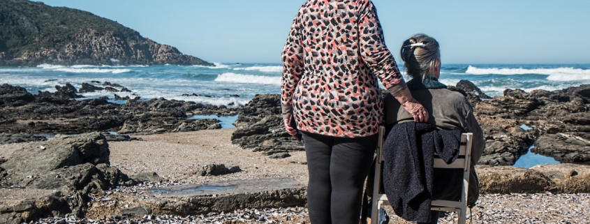 Senior woman with dementia sitting on a chair and her caregiver by the sea