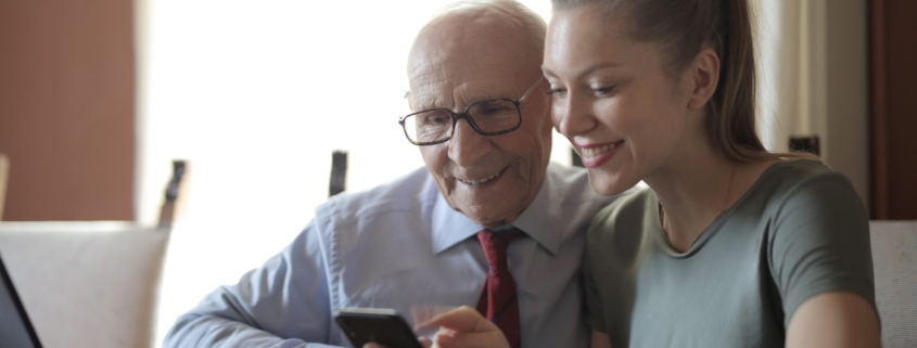 Young positive lady showing photos on smartphone to senior man while sitting at laptop